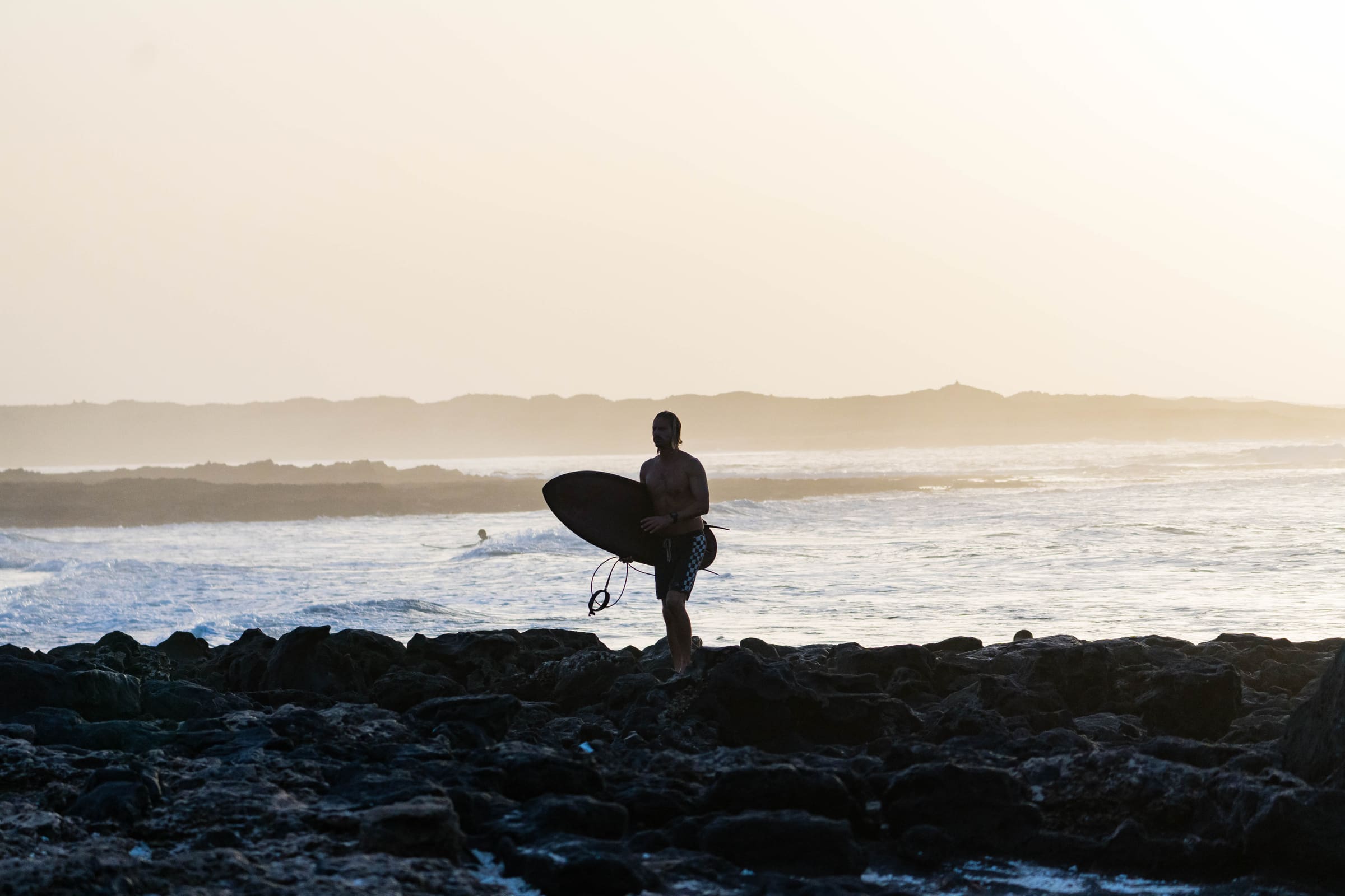 Surfeur silhouette portant sa planche sur des rochers volcaniques au coucher du soleil
