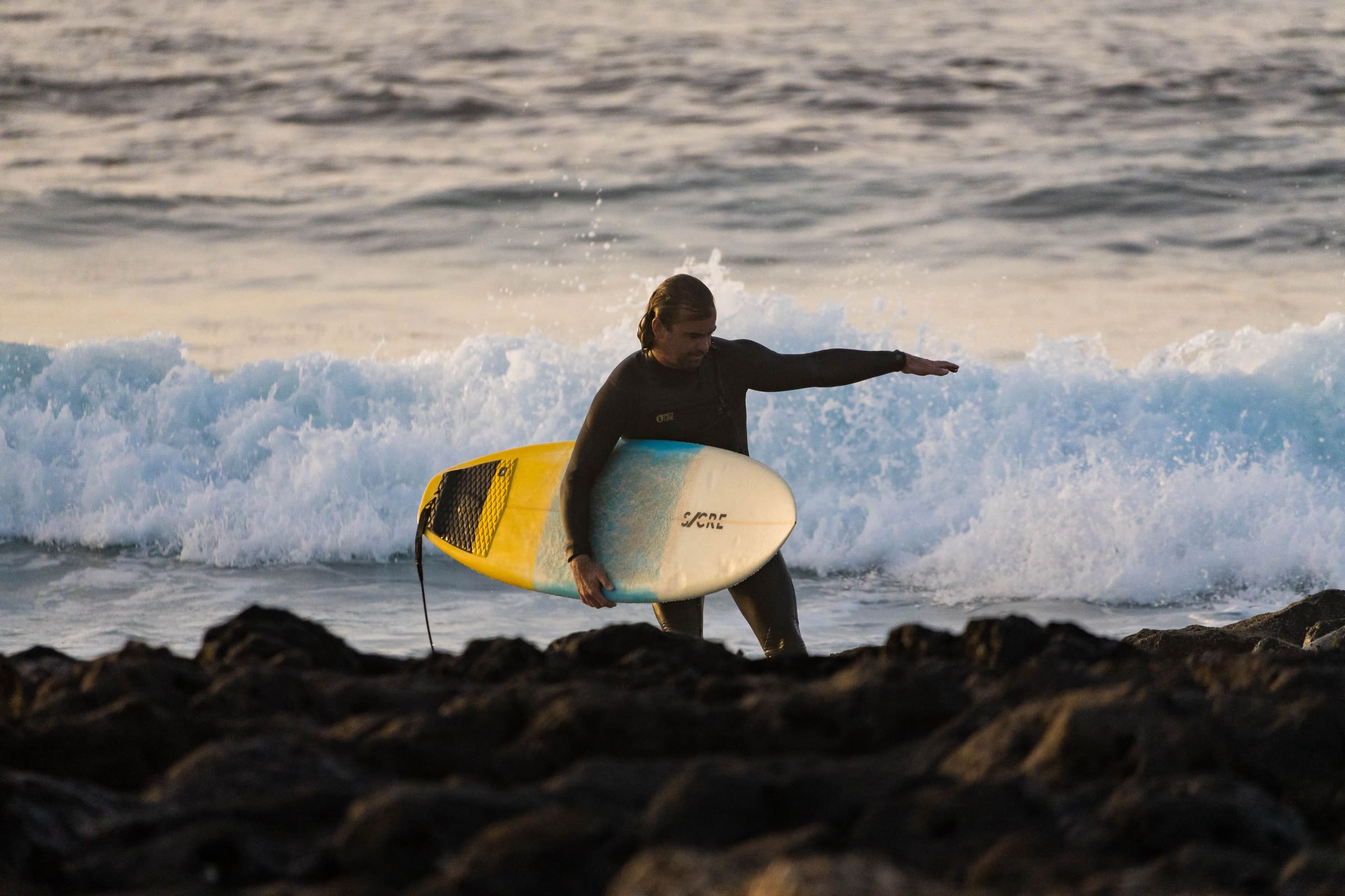 Surfeur portant sa planche jaune devant une vague qui déferle